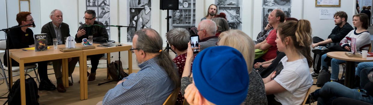 Eine Veranstaltung auf der Leipziger Buchmesse. Drei Personen sitzen an einem Tisch mit Mikrophon im Hintergrund. Im Vordergrund und im rechten Bild sitzen Besucher:innen. 