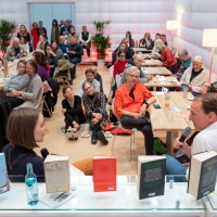 In der #buchbar sitzt im Vordergrund Marc Elsberg am Tresen und unterhält sich mit einer Frau. Dabei sitzen im Hintergrund viele verschiedene Personen an Tischen und auf dem Boden und Hören aufmerksam zu. 