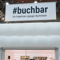 Im Eingang der #buchbar stehen mehrere Besucher, im inneren sitzen sie an den Tischen und lauschen Marc Elsberg, welcher am Tresen sitzt. Über dem Eingang hängt ein Banner mit #buchbar "Ein Projekt der Leipziger Buchmesse". 