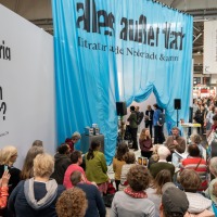 Der Stand des Gastlandes 2024 Niederlande und Flandern. Im Himtergrund hängt ein Blauer Vorhang mit der Aufschrift "alles außer flach". Im Vordergrund hören viele Menschen der Lesung zu. Der Stand des Gastlandes 2024 Niederlande und Flandern. Im Himtergrund hängt ein Blauer Vorhang mit der Aufschrift "alles außer flach". Im Vordergrund hören viele Menschen der Lesung zu.