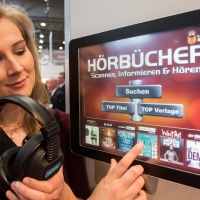 Junge Frau mit Kopfhörern in der Hand zeigt auf einen digitalen Bildschirm, der eine Bedienoberfläche mit Hörbüchern zeigt