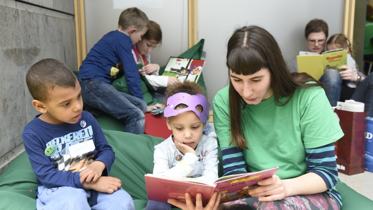 Junge Frau liest zwei Kindern aus einem Buch vor. Im Hintergrund sind weitere lesende Kinder zu sehen.