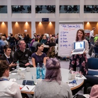Die Veranstaltung Leipziger Autor:innenrunde im Congress Center Leipzig. Viele Menschen sitzen um Runde Tische herum. Im Vordergrund steht eine Frau am Tisch und hält einen Laptop hoch. 