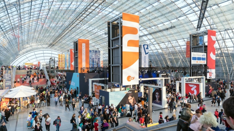 Die Glashalle vom inneren mit vielen Besucher:innen und Ständen. In der Mitte hängen viele Banner der Medien.