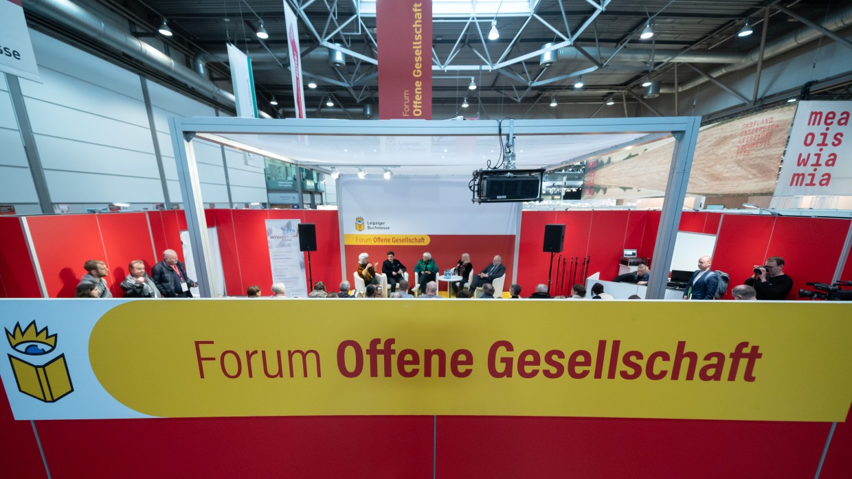 Das Forum Offene Gesellschaft der Leipziger Buchmesse von außen mit großem Schriftzug im Vordergrund. Im Hintergrund sieht man das Forum von innen während einer Diskussionsrunde.