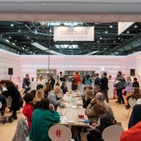 In der #buchbar sitzt das Publikum an langen Tischen während im Hintergrund Menschen an einer Bartheke zu sehen sind. An der gleichen Bar sitzen links 3 Personen mit Mikrophonen im Gespräch.