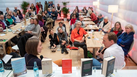 In der #buchbar sitzt im Vordergrund Marc Elsberg am Tresen und unterhält sich mit einer Frau. Dabei sitzen im Hintergrund viele verschiedene Personen an Tischen und auf dem Boden und Hören aufmerksam zu. 