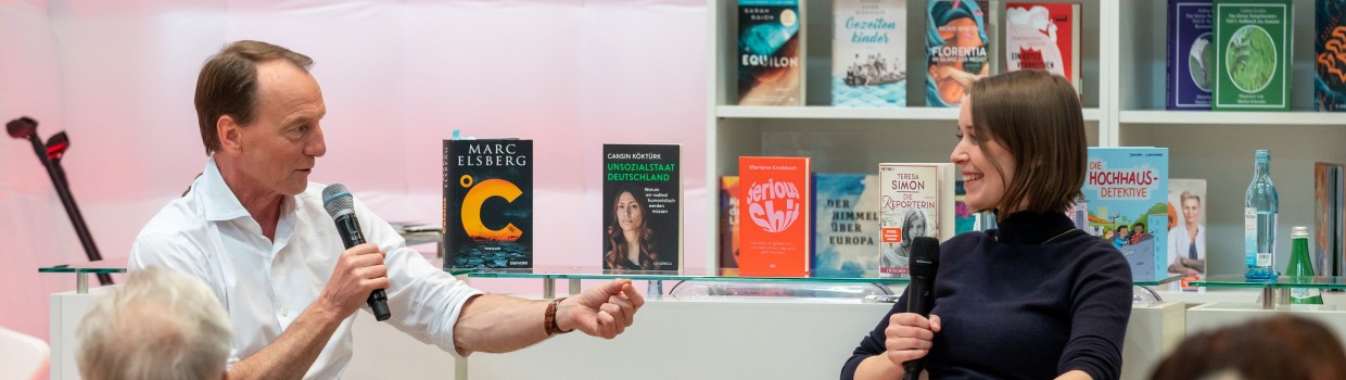 In der #buchbar sitzt Marc Elsberg am Tresen und unterhält sich mit einer Frau. Dabei sitzen im Vordergrund Personen an Tischen und hören zu. Im Hintergrund sieht man Tresen und Regale, gefüllt mit Büchern.