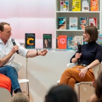 In der #buchbar sitzt Marc Elsberg am Tresen und unterhält sich mit einer Frau. Dabei sitzen im Vordergrund Personen an Tischen und hören zu. Im Hintergrund sieht man Tresen und Regale, gefüllt mit Büchern.