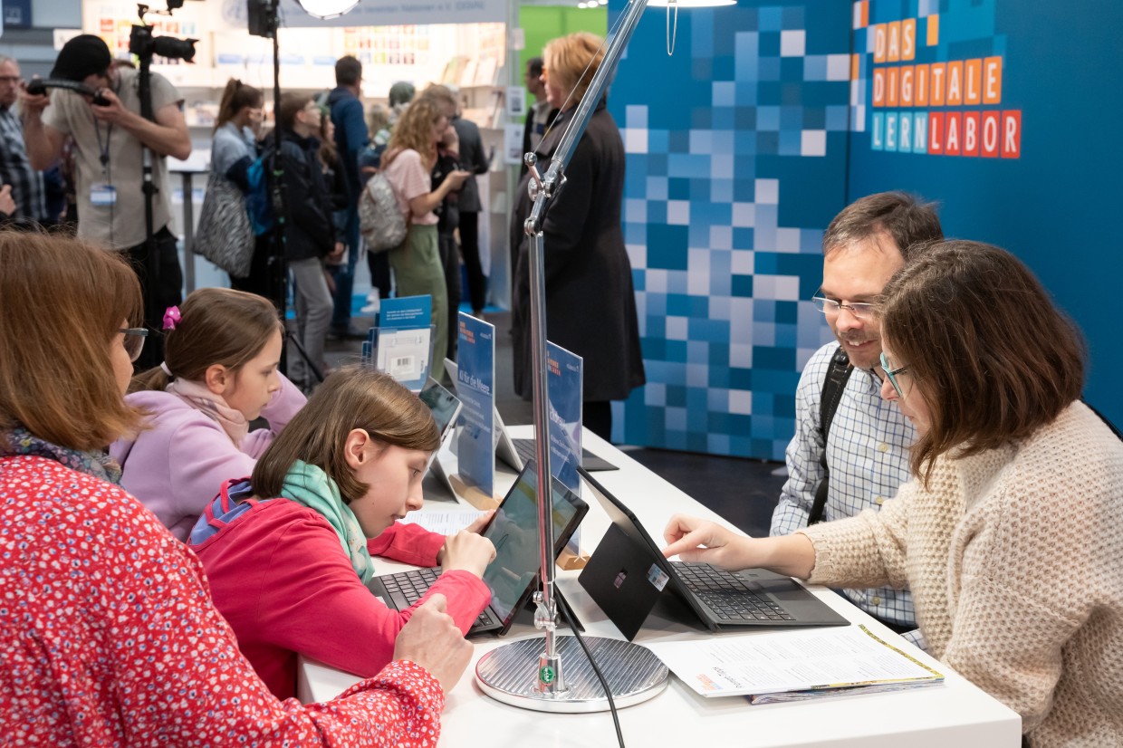 Im Digitalen Lernlabor der Leipziger Buchmesse 2023 sitzen Kinder und Erwachsene an Laptops, im Hintergrund stehen Besucher:innen