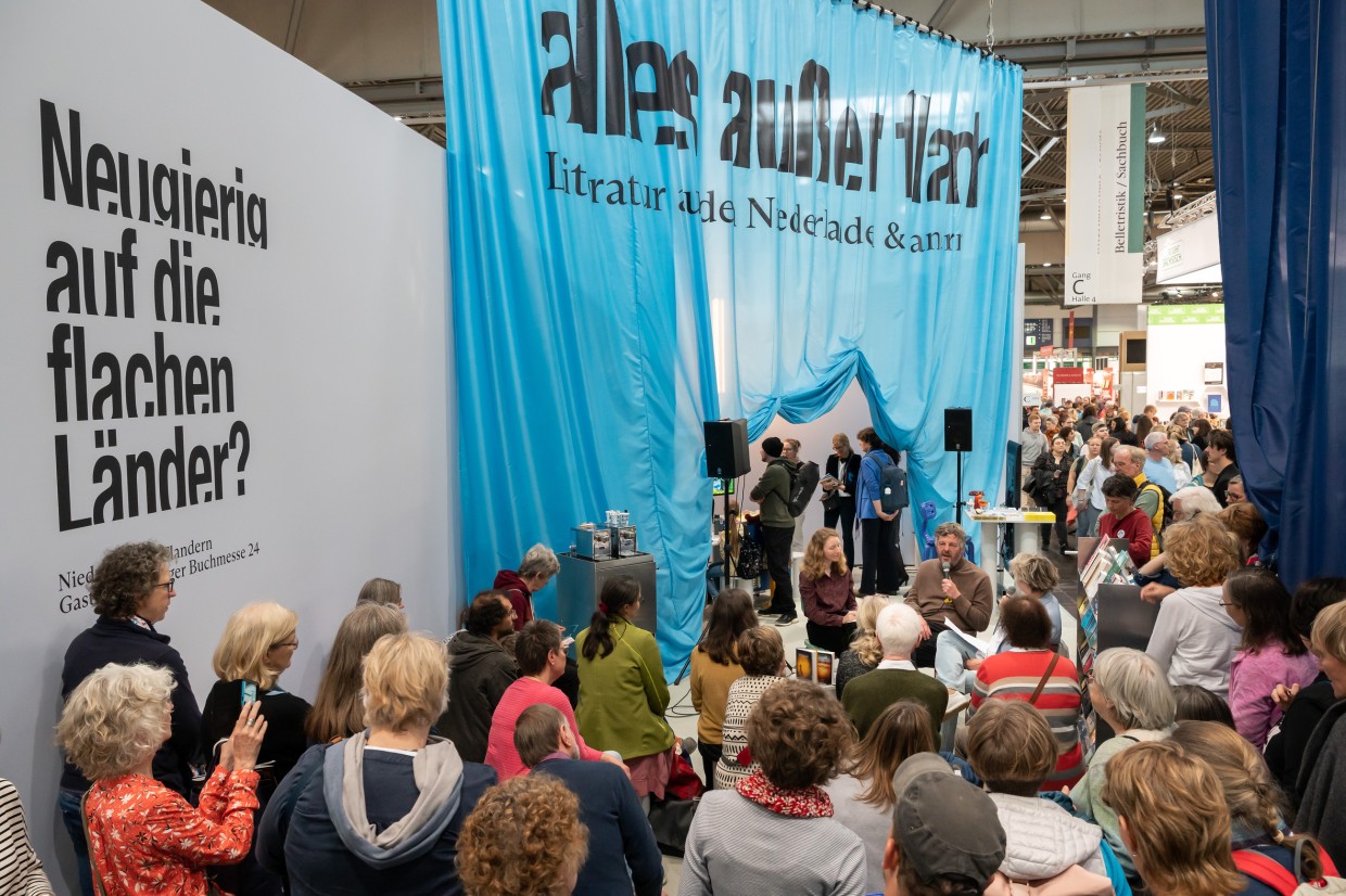 Der Stand des Gastlandes 2024 Niederlande und Flandern. Im Himtergrund hängt ein Blauer Vorhang mit der Aufschrift "alles außer flach". Im Vordergrund hören viele Menschen der Lesung zu.