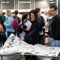 Besucher:innen blättern in einem riesigen illustrierten Buch im Buch- und Druckkunsbereich auf der Leipziger Buchmesse 2023