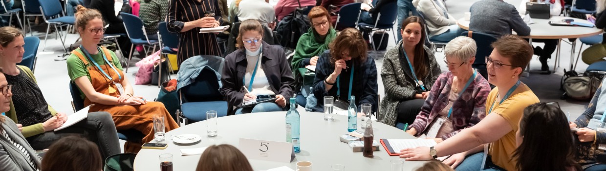 Vollbesetzter Saal der Leipziger Autor:innenrunde 2024 mit einem großen runden Tisch im Vordergrund, an dem sich Teilnehmerinnen austauschen