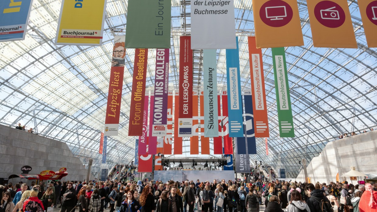 Innenaufnahme der Glashalle der Leipziger Messe mit bunten Werbebannern und vielen Besucher:innen während der Leipziger Buchmesse 2024 Innenaufnahme der Glashalle der Leipziger Messe mit bunten Werbebannern und vielen Besucher:innen während der Leipziger Buchmesse 2024