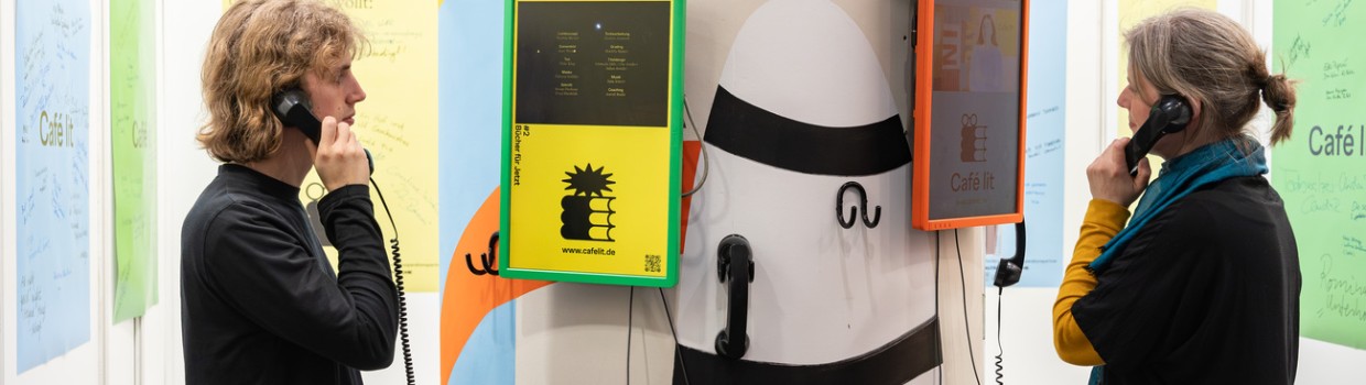 Messestand von Café lit auf der Leipziger Buchmesse 2024 mit 2 Personen, die an einer Litfaßsäule telefonieren.