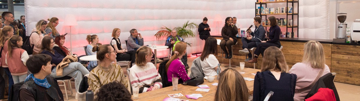 Eine Autorin im Gespräch mit den beiden Moderator:innen in der #buchbar während der Leipziger Buchmesse 2025 vor vollbesetztem Publikum