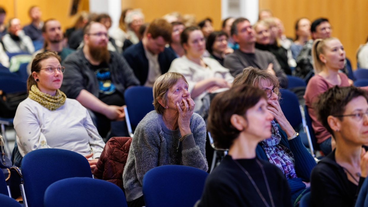 vollbesetztes Publikum in einem großen Raum hört aufmerksam zu