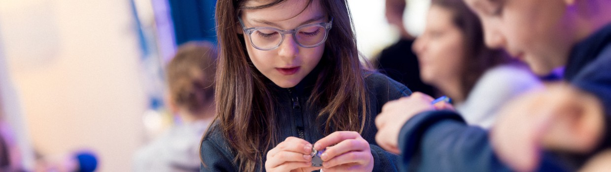 mehrere Kinder sitzen an einem Tisch, ein Mädchen betrachtet interessiert ein Objekt in ihren Händen während der Leipziger Buchmesse 2025