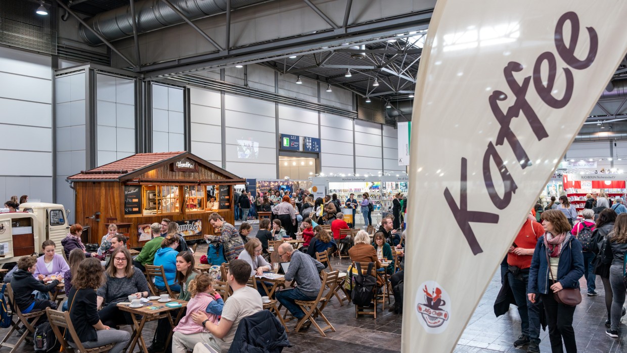 Einer der gastronomischen Bereiche in der Messehalle der Leiziger Buchmesse 2025 mit vielen Besucher:innen an Tischen und einem Verkaufsstand im Hintergrund