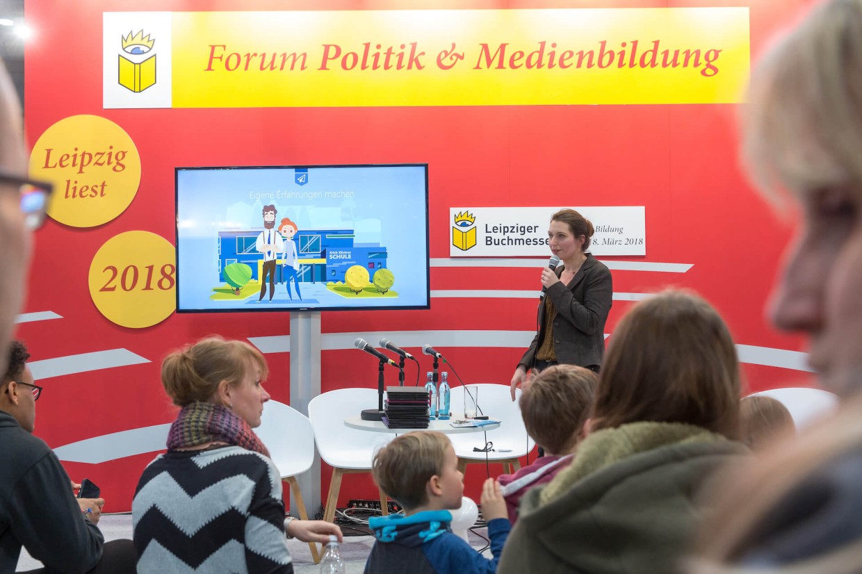 Eine Frau steht neben einem Fernseher und spricht ins Mikrofon. Davor sitzen Menschen und hören ihr zu. Im Hintergrund eine rote Aufstellwand mit der Aufschrift "Forum Politik und Medienbildung". 