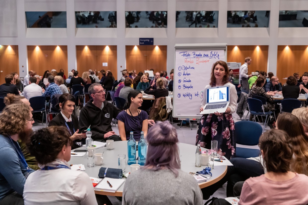 Die Veranstaltung Leipziger Autor:innenrunde im Congress Center Leipzig. Viele Menschen sitzen um Runde Tische herum. Im Vordergrund steht eine Frau am Tisch und hält einen Laptop hoch. 