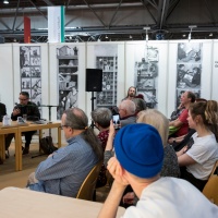 Eine Veranstaltung auf der Leipziger Buchmesse. Drei Personen sitzen an einem Tisch mit Mikrophon im Hintergrund. Im Vordergrund und im rechten Bild sitzen Besucher:innen.  Eine Veranstaltung auf der Leipziger Buchmesse. Drei Personen sitzen an einem Tisch mit Mikrophon im Hintergrund. Im Vordergrund und im rechten Bild sitzen Besucher:innen.