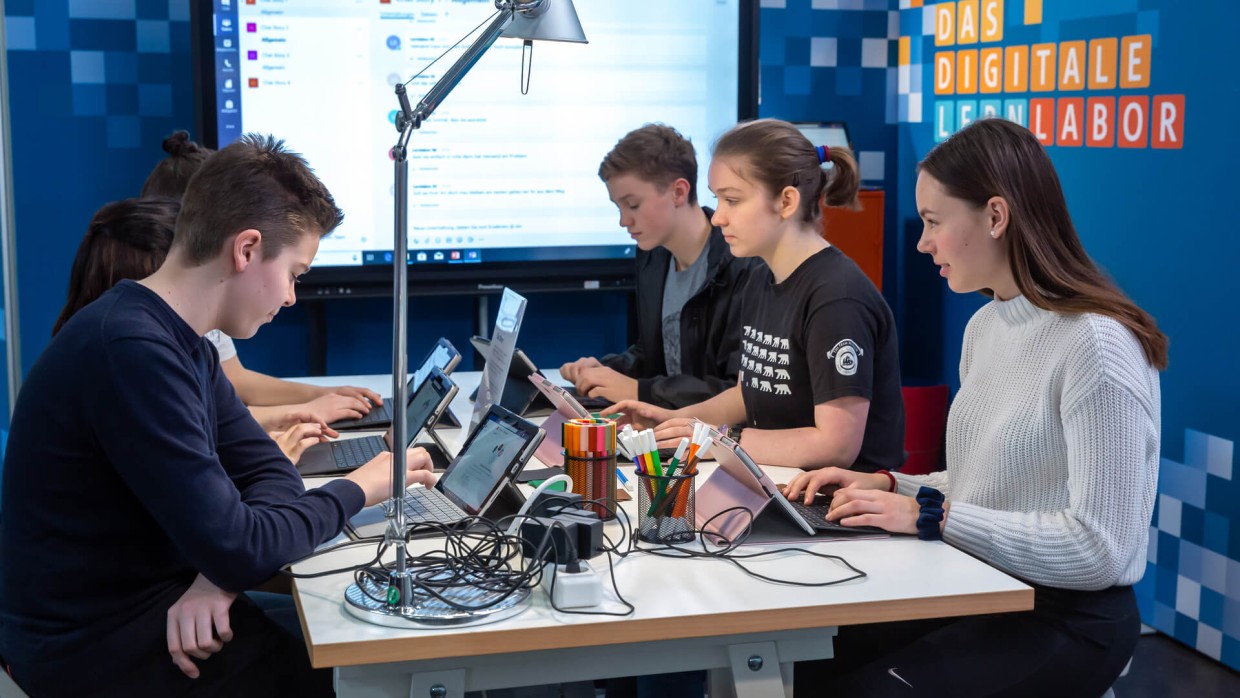 Im digitalen Lernlabor auf der Leipziger Buchmesse. Sechs junge Jugendliche sitzen sich gegenüber und haben ein Surface aufgeklappt. Im Hintergrund ist ein großer Monitor zu sehen.