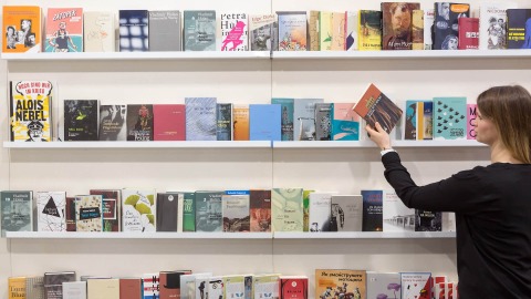 A woman is standing in front of a bookshelf and reaching for a book. The woman is dressed in black and is standing on the right side of the picture. The bookshelf consists of four white shelves. 