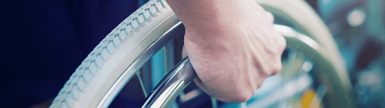 Ein Foto von einem Ausschnitt eines Rollstuhls. Zu sehen ist der obere Teil eines Rades und eine Hand die danach greift. 