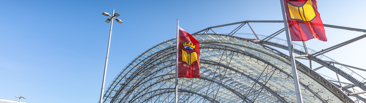 Auf dem Foto ist die Glashalle von außen zu sehen. Der Himmel ist blau und an den Fahnenstangen hängen Flaggen mit dem Buchmesse-Logo. 