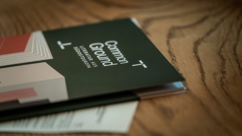 Auf einem Holztisch liegt ein dunkelgrünes Heft mit dem Titel „Common Ground – Literatur aus Südosteuropa“. Darunter steckt ein weiteres Blatt Papier.
