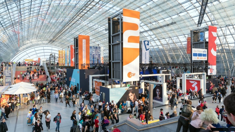 The glass hall from the inside with many visitors and stands. In the middle, there are many media banners hanging.