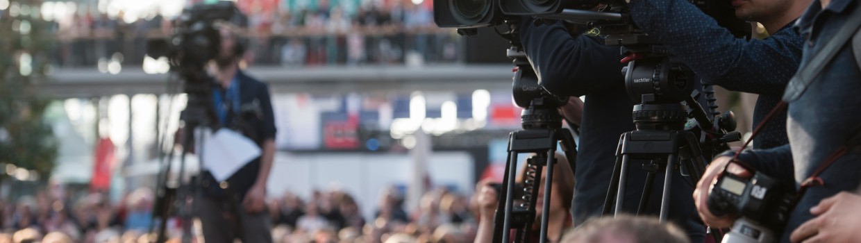 A photo from an event at the Leipzig Book Fair. Three cameramen and their cameras can be seen pointing at an object outside the frame. Two of the men are clearly visible on the right side of the image. One is blurred in the background. 