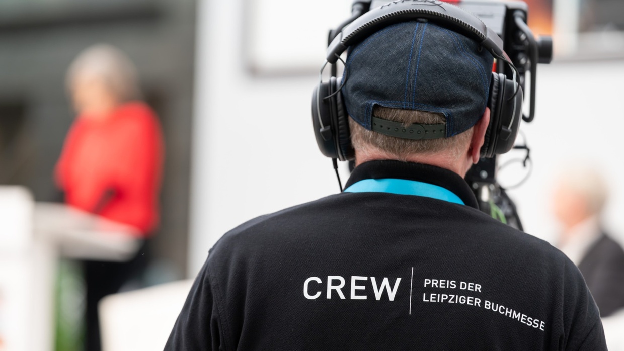 Cameraman from behind wearing large headphones and a T-shirt with the word “Crew” on it, filming the award ceremony during the Leipzig Book Fair 2024 with a large camera