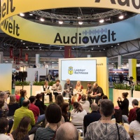 Stage event with several people in the audio world of the 2025 Leipzig Book Fair in front of a full audience.
