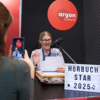 A woman is speaking a text into a microphone at the booth of the exhibitor Argon Hörbuch in the audio world of the 2025 Leipzig Book Fair, while another woman is filming her with a smartphone.
