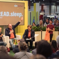 Stage event of the podcast "eat.read.sleep." with several people in the audio world of the 2025 Leipzig Book Fair in front of a full audience
