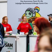 several members of the Awareness Team SAM of the Leipzig Book Fair 2025 are standing at the booth while visitors walk past
