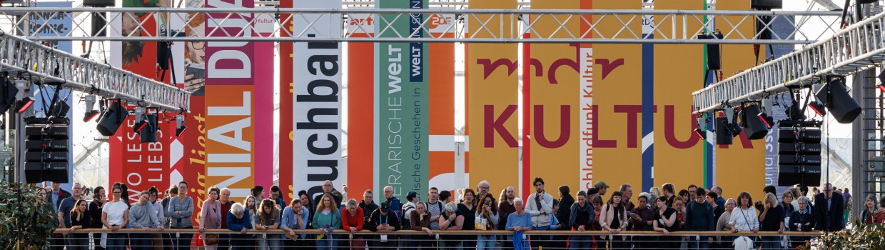Many people are standing on the bridge in the glass hall of the exhibition center. Behind them hang many flags.
